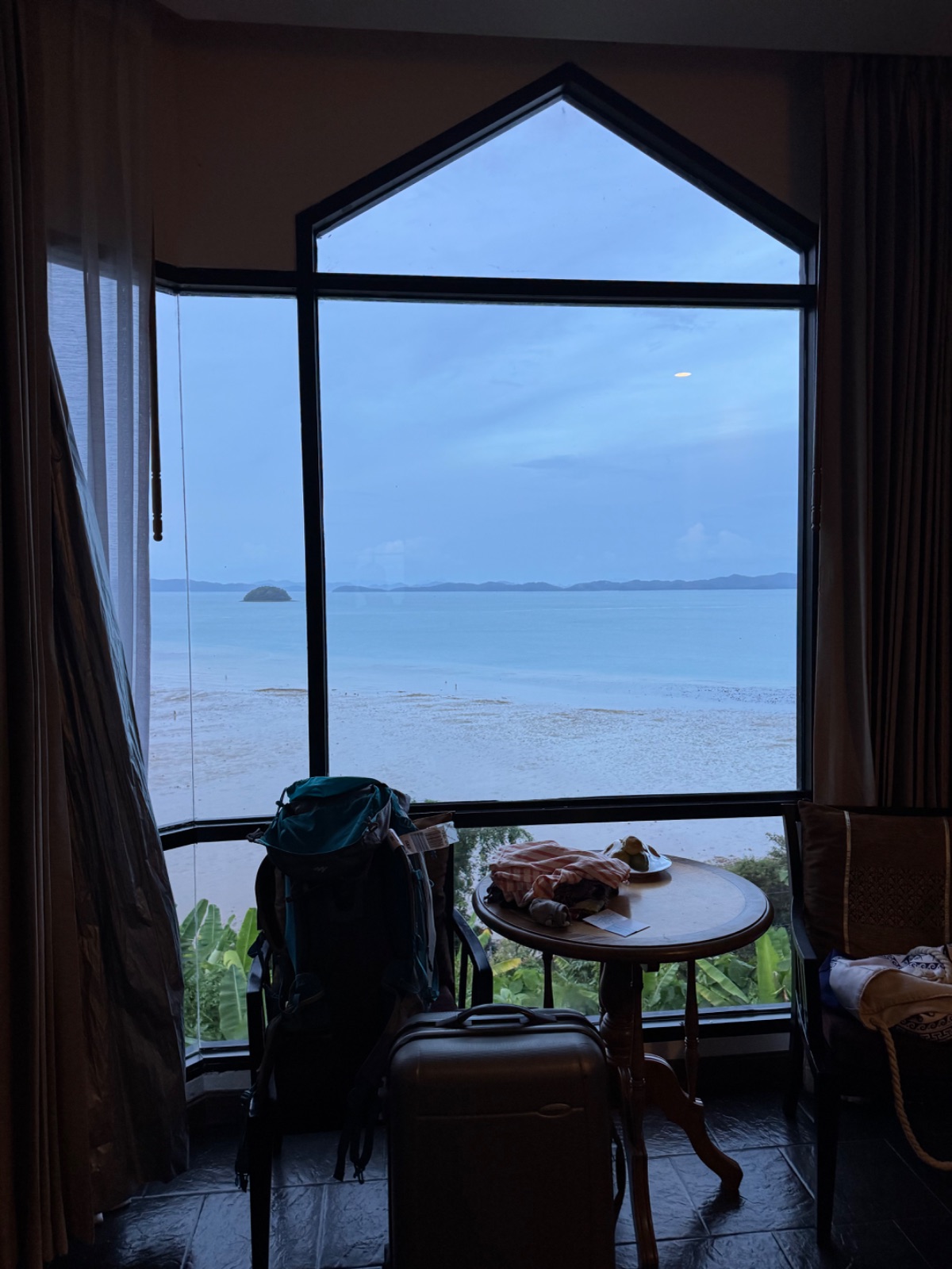 Grande fenêtre cathédrale de la chambre d'hôtel avec vue sur la mer à marée basse, sacs de voyage au premier plan