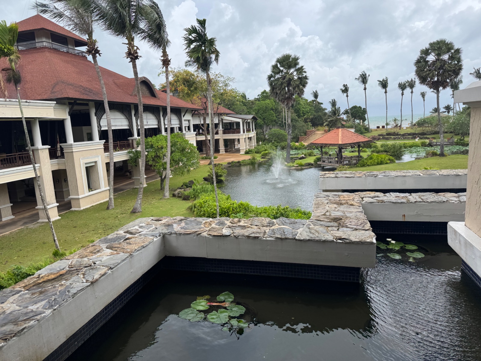 Jardins du Rawi Warin Resort à Koh Lanta : étang avec fontaine, palmiers, pavillons et mer en arrière-plan